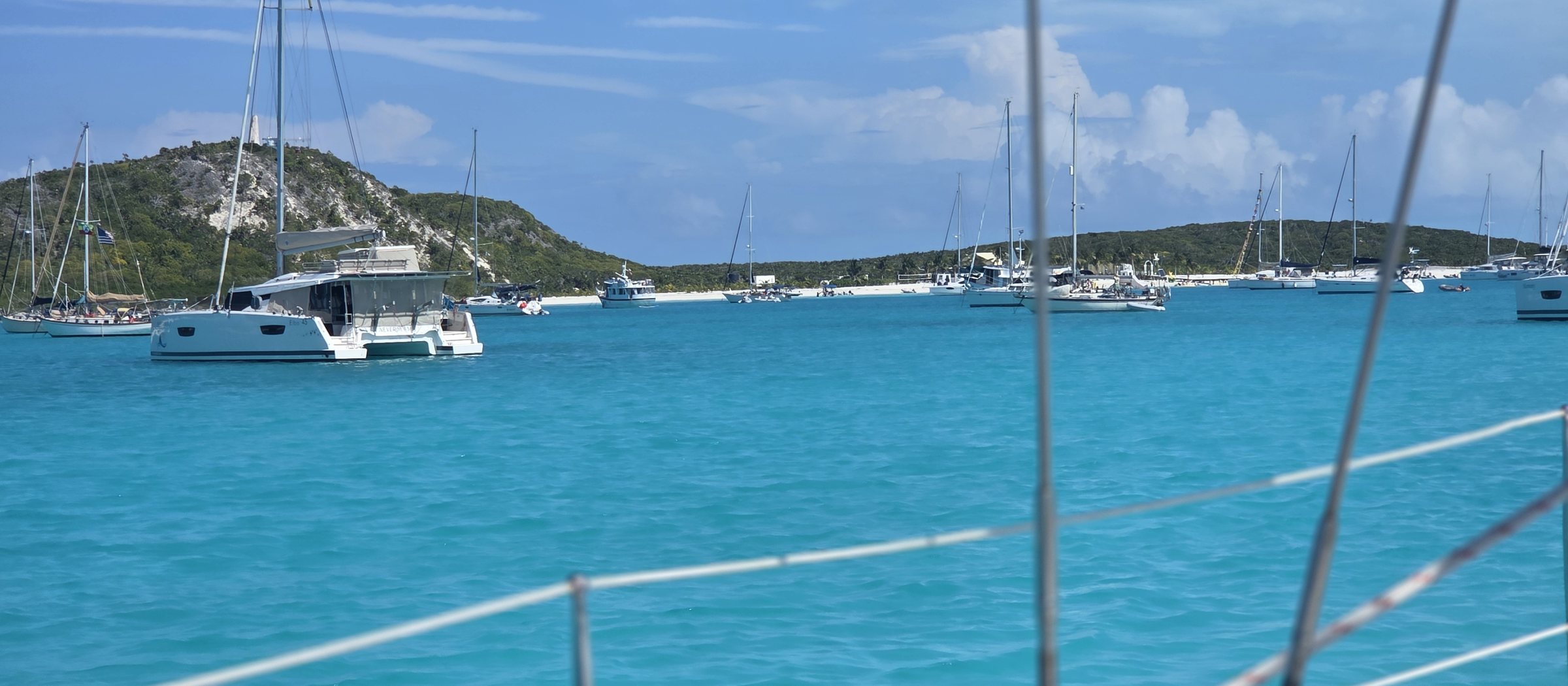 Turquoise anchorage in the Bahamas seen through SV Salty's lifelines, with sailboats and a catamaran at anchor below green hills.