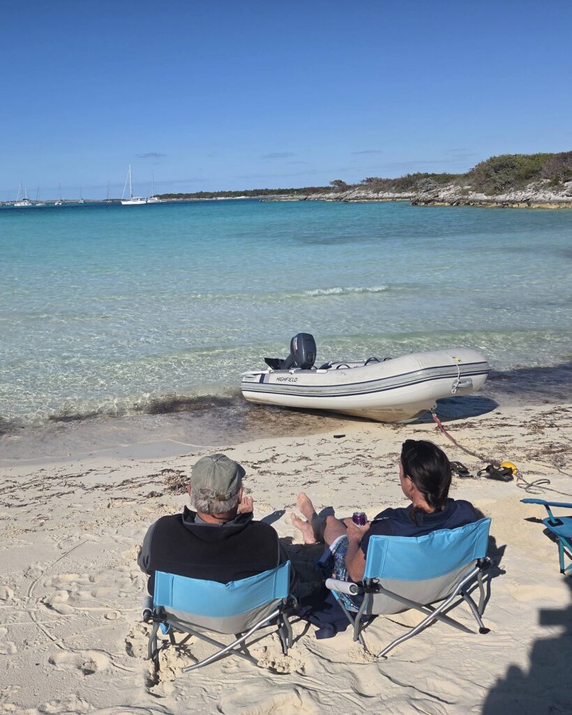 Two people relax in blue folding beach chairs on white sand, facing clear turquoise water where a gray inflatable dinghy sits on the beach under a deep blue sky.