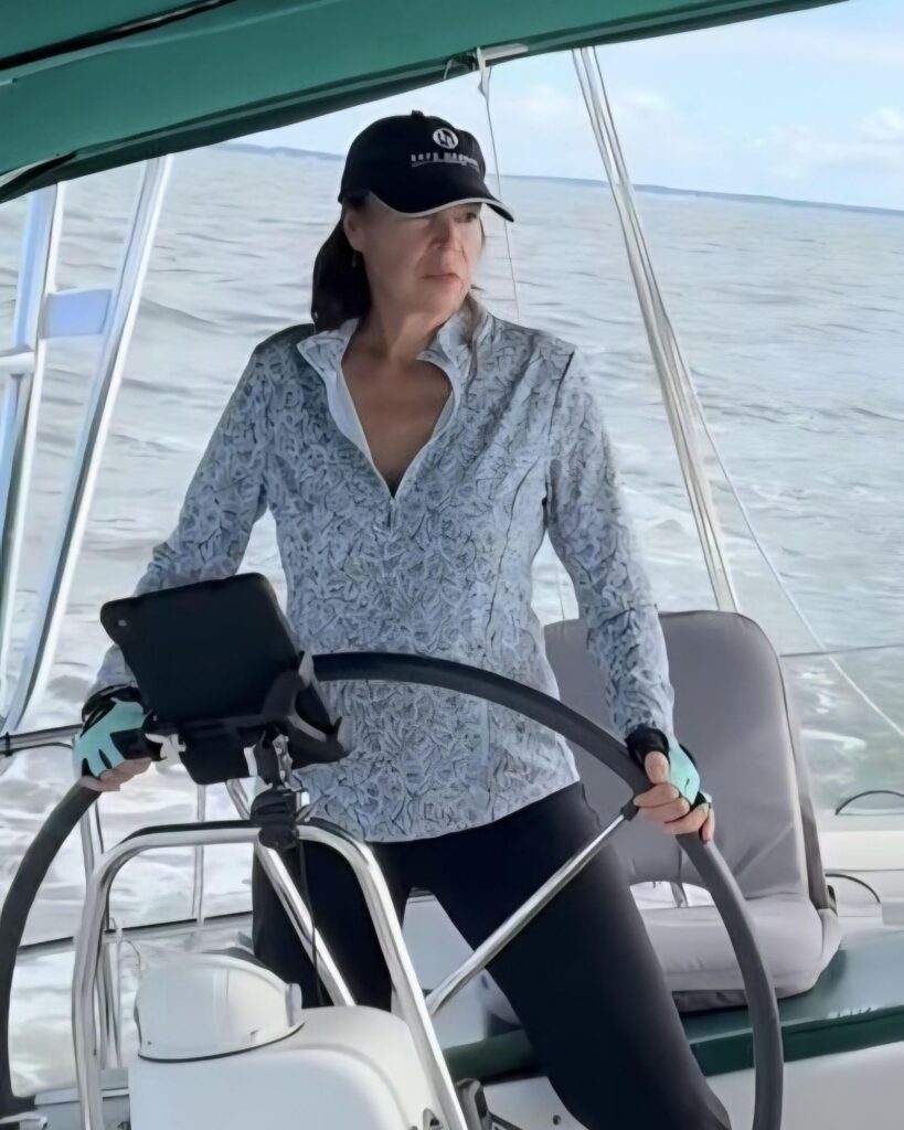Beth at SV Salty's helm, in a dark cap and patterned top, steering at the wheel under the green bimini.