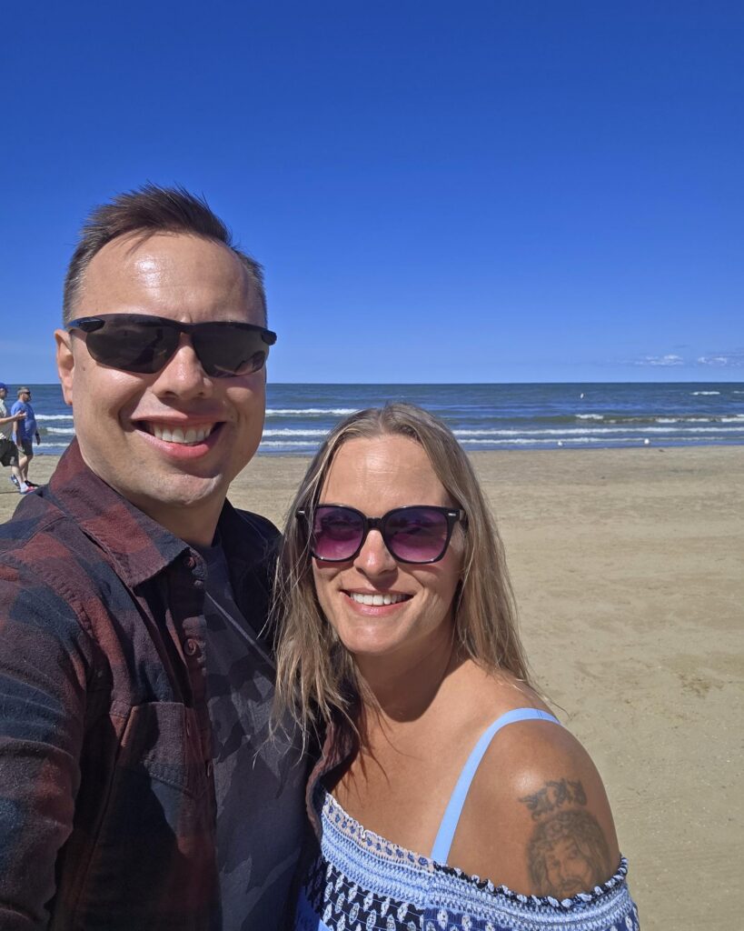 Brad and Kelley smile for a selfie on a sandy beach in Cleveland, Ohio, both in sunglasses, Brad in a plaid shirt and Kelley in a blue patterned top, with waves and a deep blue sky behind them.