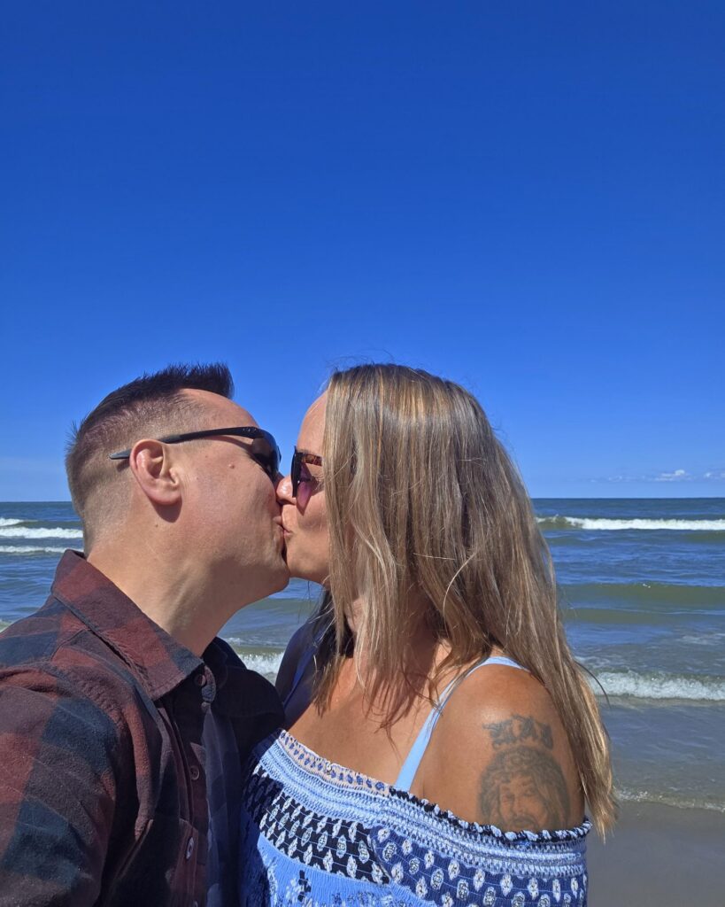 Brad and Kelley share a kiss on the beach in Cleveland, Ohio, in sunglasses, with breaking waves and a clear blue sky behind them.
