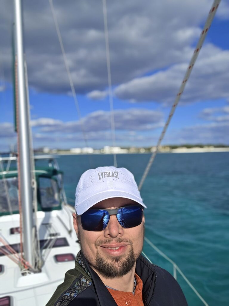 Brad smiles in a white Everlast cap and mirrored sunglasses at the bow of SV Salty, with turquoise water, a distant shoreline and a cloudy blue sky behind him.