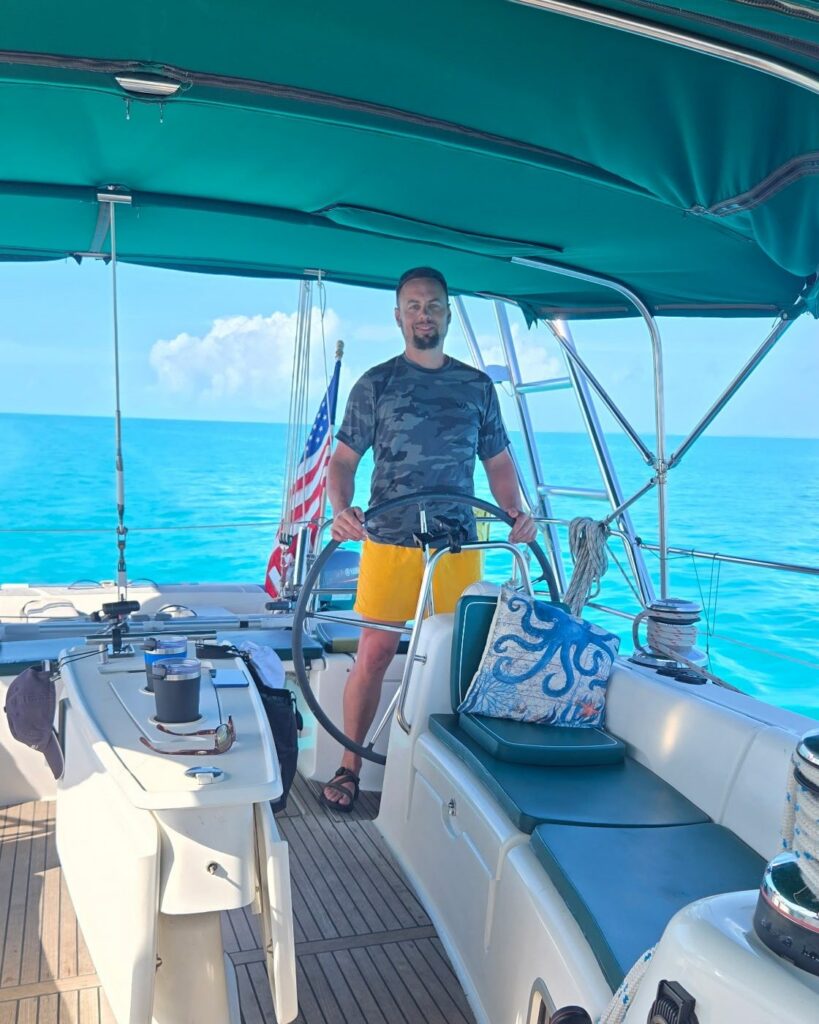 Brad standing at Salty's wheel in a camo T-shirt and yellow shorts, under the teal bimini, with bright turquoise water all around.