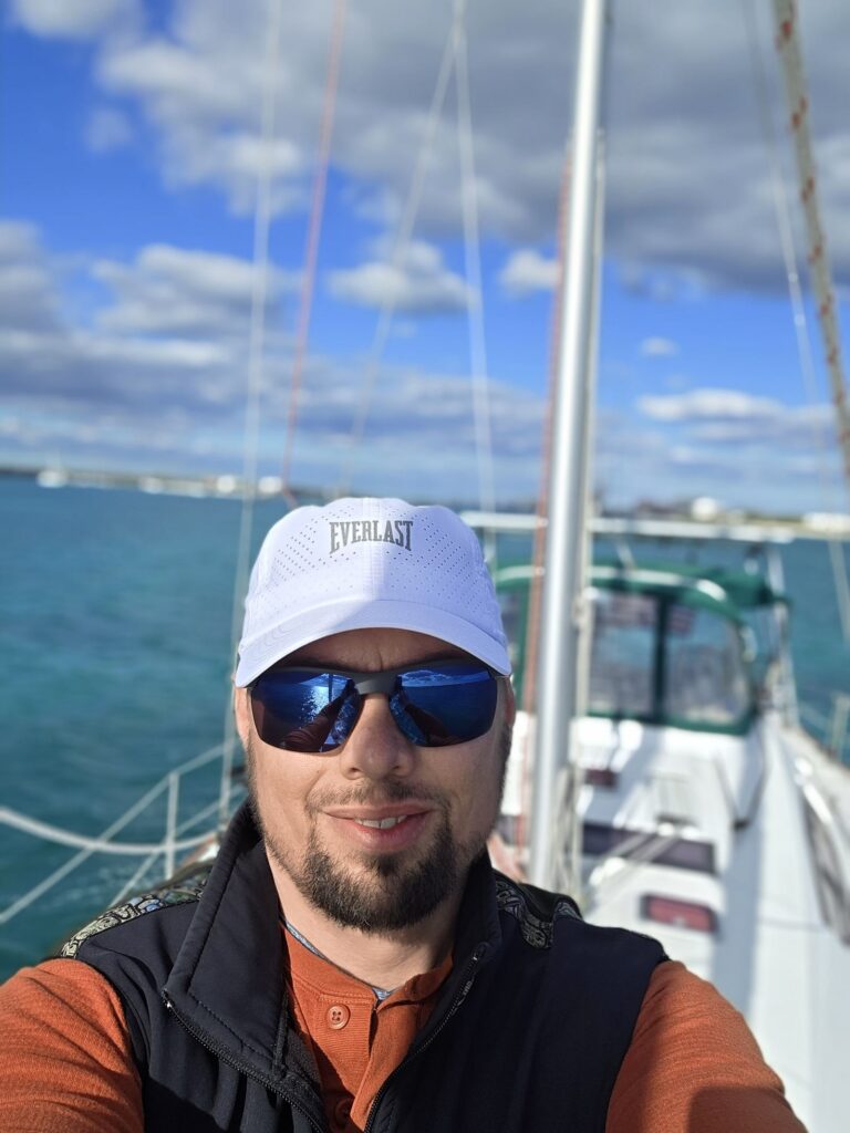 Brad takes a selfie on SV Salty's foredeck in a white Everlast cap, sunglasses and a dark vest, with the mast, green-trimmed dodger and turquoise water behind him.
