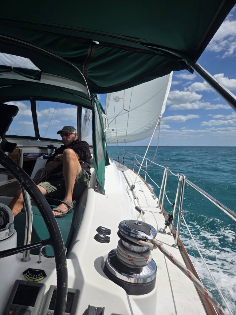 A man in a cap and sunglasses sits in SV Salty's cockpit under the green bimini as she sails, with the headsail full, a stainless winch in the foreground and white wake on deep blue-green water.