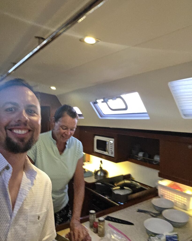 Two of the crew smiling in SV Salty's galley while dinner cooks on the stove, with the microwave and portlights behind them.