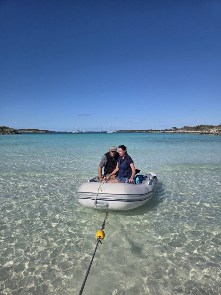 Two people sit in a gray inflatable dinghy floating in crystal-clear shallow water over white sand, tied to a yellow float, with a low island under a deep blue sky.