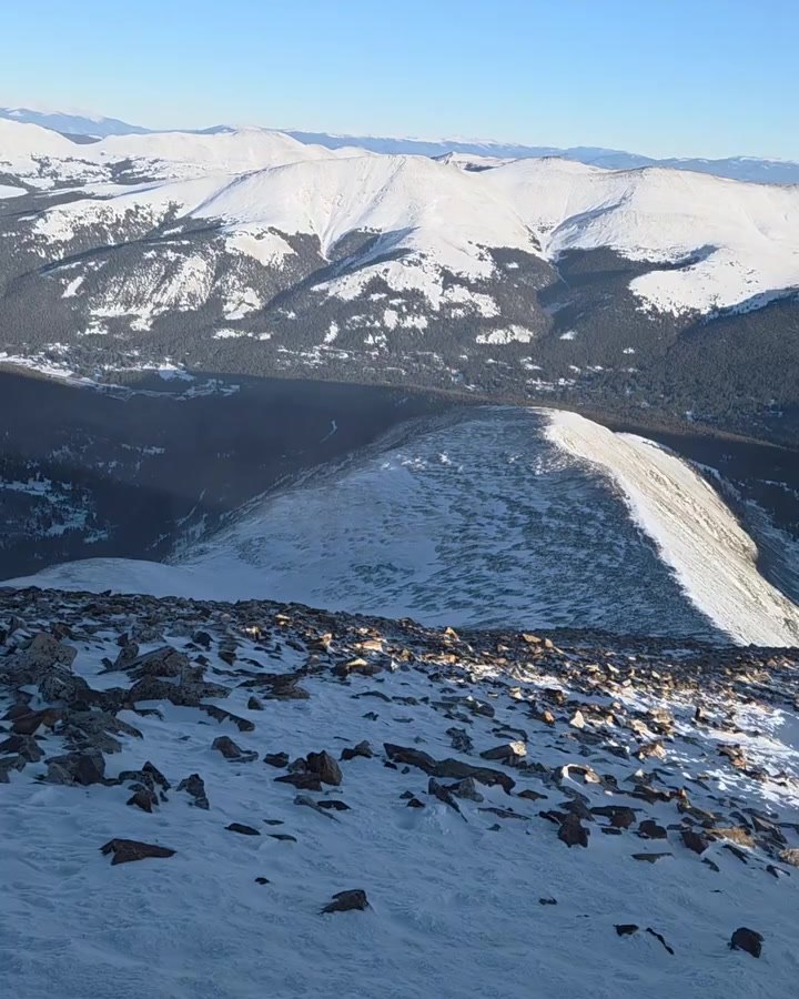 View from a rocky, snow-covered summit across a snowy ridgeline to rolling white mountains under a clear blue sky