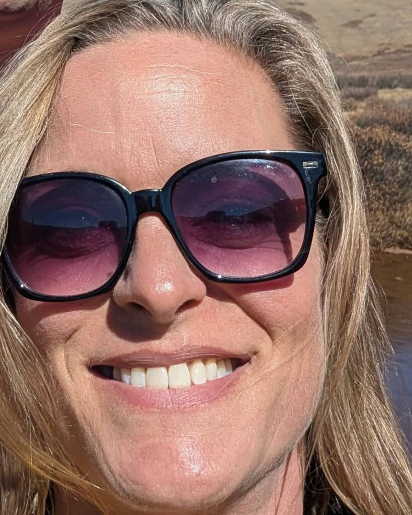 Kelley smiling in pink-tinted sunglasses, her long blond hair framing her face, on a sunny day outdoors.