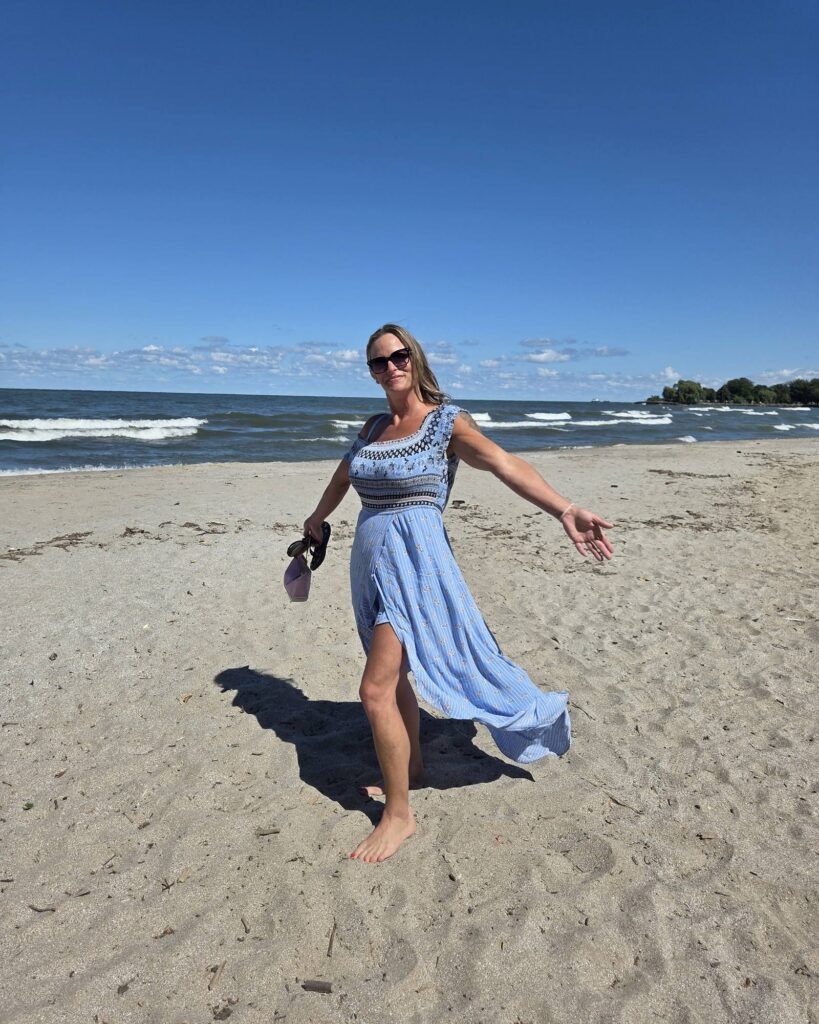 Kelley twirls barefoot on the sand in Cleveland, Ohio, in a flowing light blue dress and sunglasses, sandals in hand, with waves, a wooded point and a blue sky behind her.