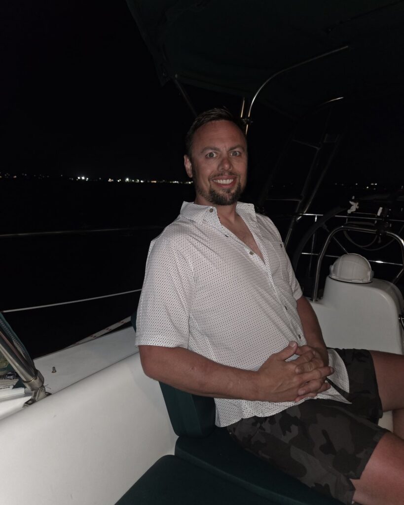 A crew member in a white shirt relaxing in SV Salty's cockpit at night, with anchorage lights on the dark water behind.