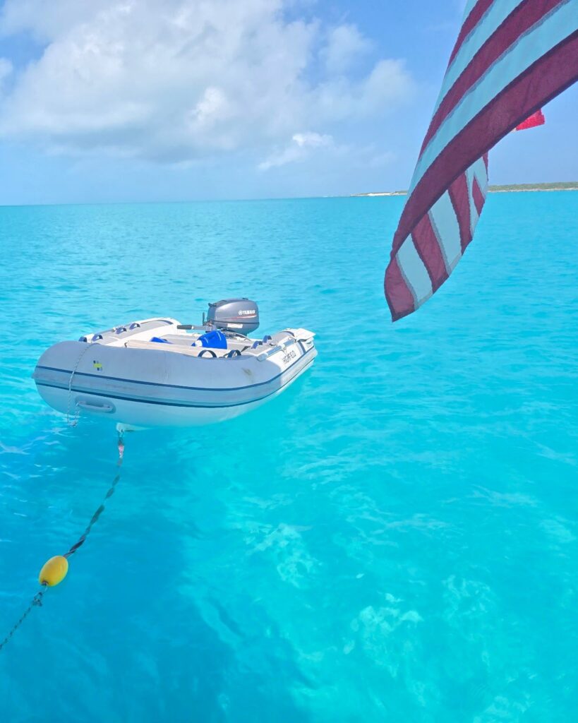 Salty's gray inflatable dinghy Pepper with its outboard, tied off the stern in clear turquoise water, with the US flag in the corner of the frame.