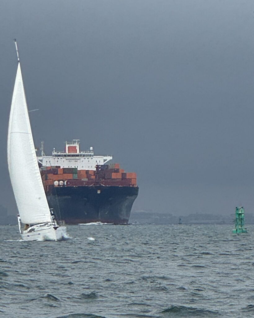SV Salty under a full white headsail sails through choppy gray water in front of a large container ship stacked with red and orange containers, under a dark storm sky, with a green channel buoy off to the right.