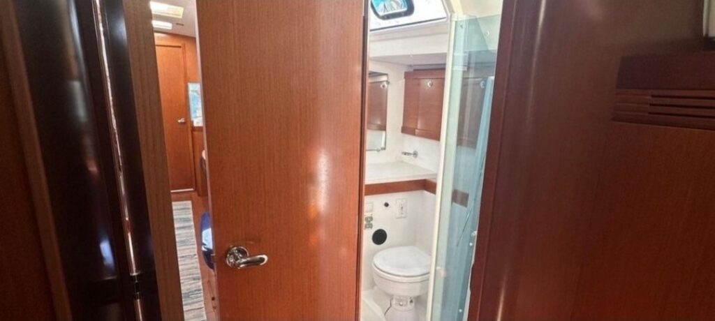 Through an open wooden door: a marine head with a toilet, a vanity with a mirror, and a frosted glass shower stall beside it.