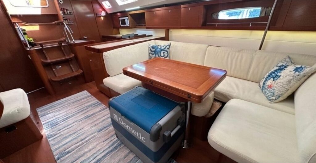 Cream L-shaped settee with octopus and seahorse pillows around a fold-out wooden table, a blue Dometic cooler, and the companionway steps and galley in the background.