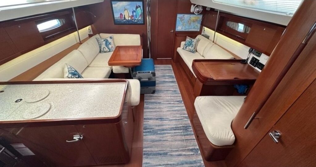 Wood-paneled sailboat salon seen from the companionway: L-shaped cream settee and table on the left, straight settee and nav desk on the right, blue striped runner down the middle.