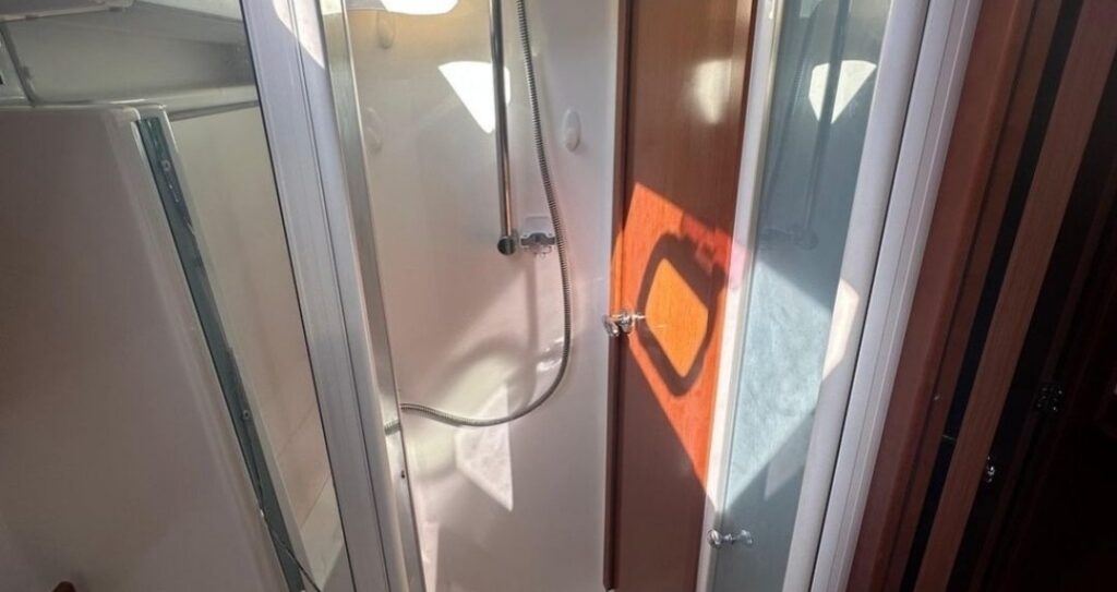 Inside a sailboat head: a white shower stall with a handheld shower on a rail and glass doors, with sunlight through a port on the wood door.