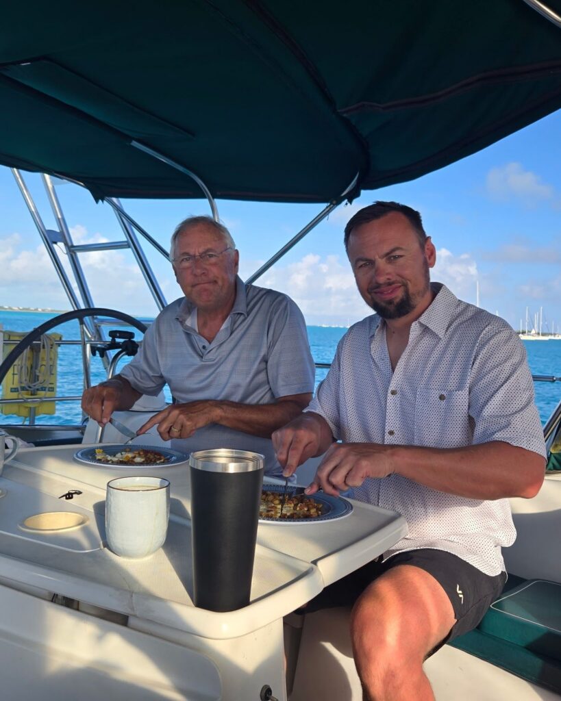 Scott and Brad eating dinner at Salty's cockpit table under the green bimini, with the wheel and an anchorage of turquoise water and sailboats behind them.