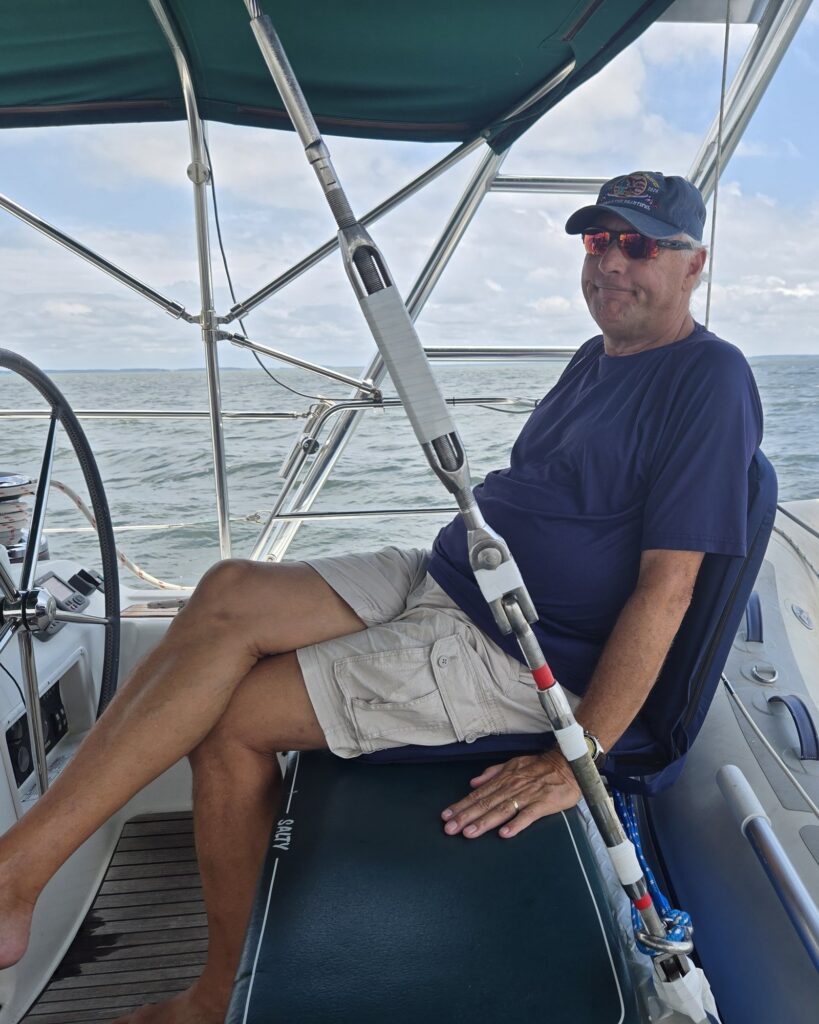 Scott, SV Salty's captain, at the main helm station in the starboard aft cockpit under the bimini, with open water behind him.