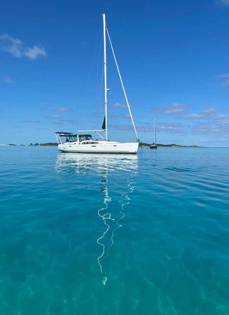 SV Salty, a white sloop with a green bimini, sits at anchor in clear turquoise water under a deep blue sky, her mast reflected in a long ripple across the calm surface.