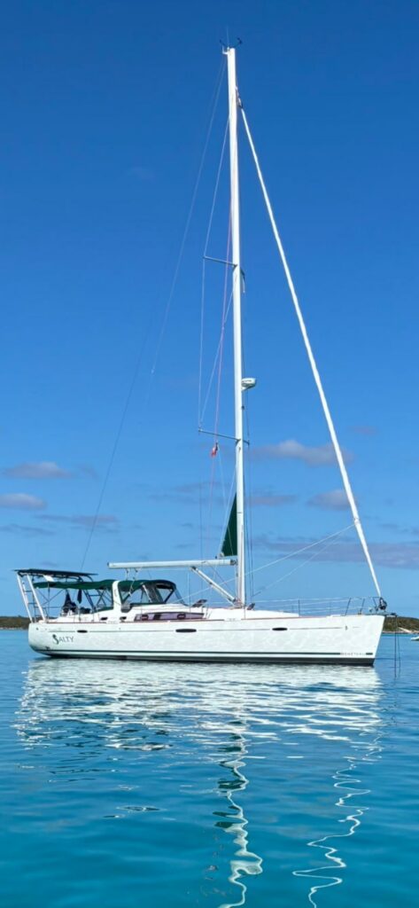 Full-length side view of SV Salty at anchor, a white sloop with SALTY and a seahorse logo on her hull, a tall mast, green bimini and dodger, and her reflection in calm blue water.