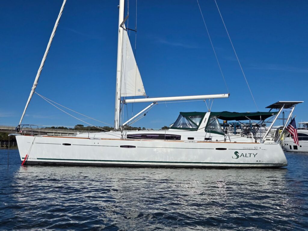 Broadside view of SV Salty under a deep blue sky, showing her Beneteau and Oceanis 50 hull marks, green canvas, the seahorse SALTY logo and a US flag at the stern.