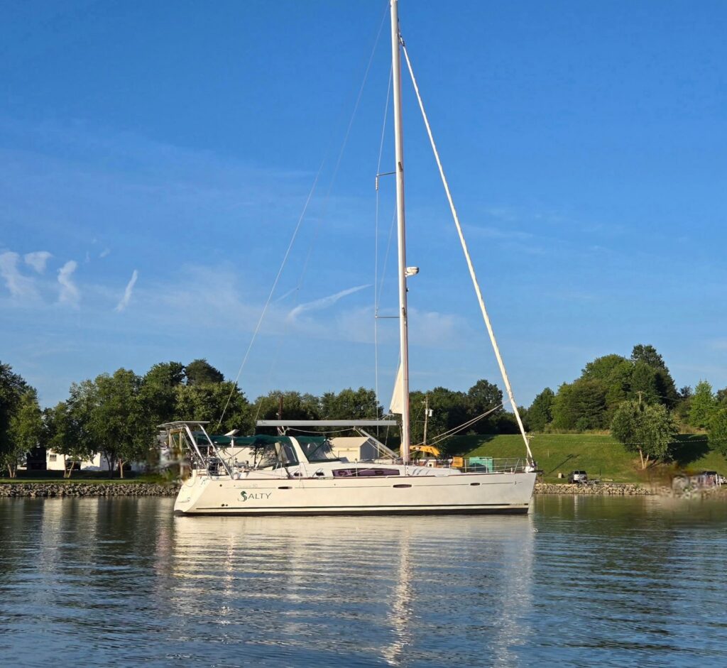 SV Salty floats on calm, glassy water off a green grassy shore lined with trees, her hull reflected below a clear blue sky.