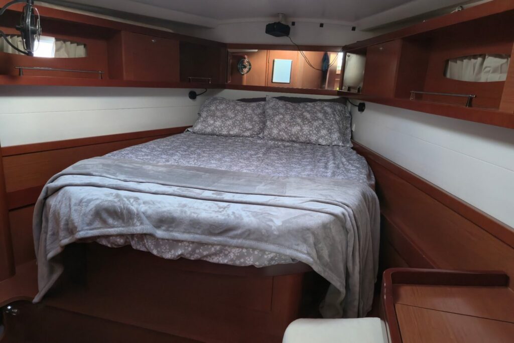 Forward master cabin with an island double berth made up in gray floral bedding and a soft gray throw, wood cabinetry and shelves on both sides, and cabin portlights above.
