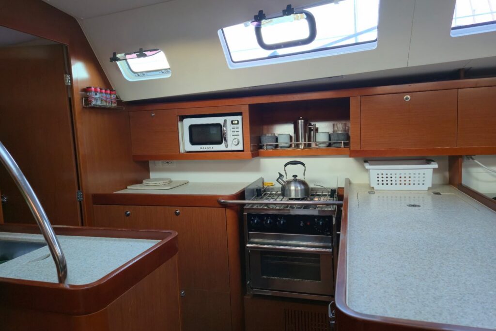 Sailboat galley with warm wood cabinets, a stainless gimbaled stove and oven with a kettle, a microwave in an upper cabinet, speckled counters on both sides, and portlights above.