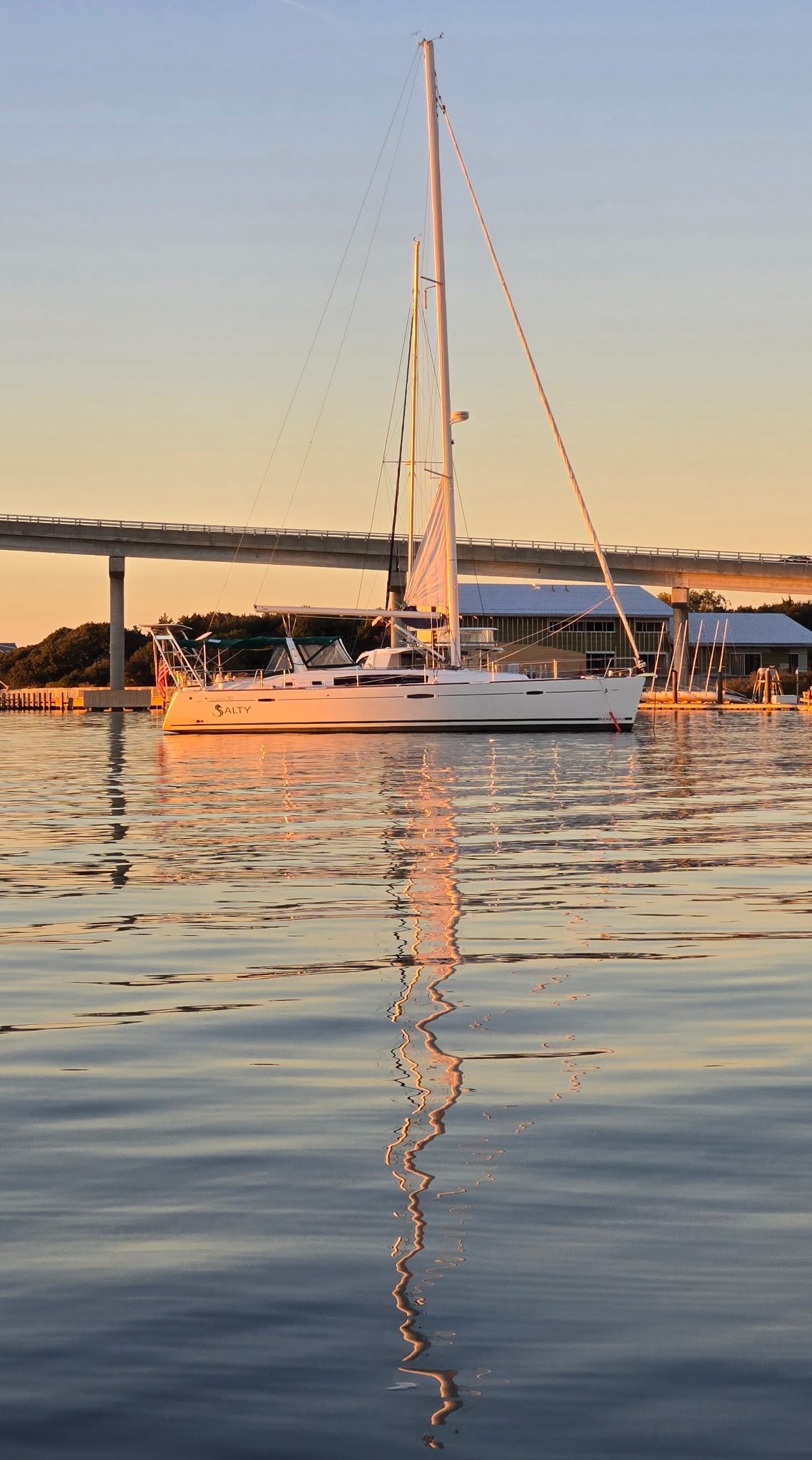 SV Salty at a dock at golden hour, her white hull and tall mast lit by the setting sun and reflected in a long zigzag across calm water, with a concrete bridge behind her.