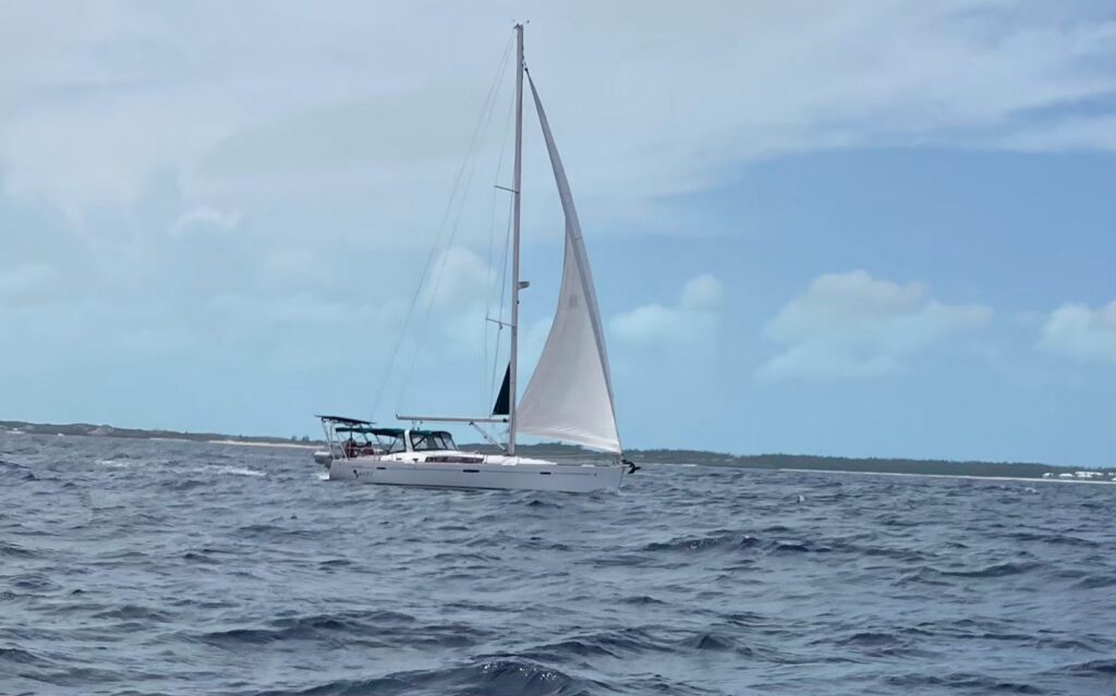 SV Salty sails under main and genoa across choppy blue water, seen from another boat, with a low wooded shoreline and soft clouds behind her.