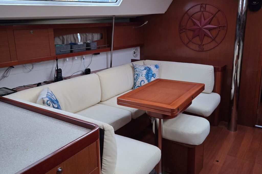 Cream L-shaped salon settee with blue sea-life pillows around a cherry table, a large compass rose on the bulkhead, and overhead hatches letting in daylight.