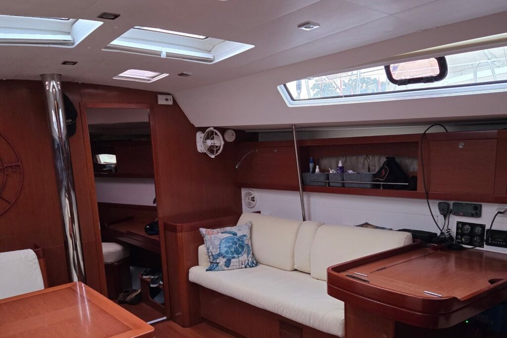 Starboard side of Salty's salon with a cream straight settee and a sea-turtle pillow, the navigation station with instrument panels at right, overhead hatches and a long hull window.