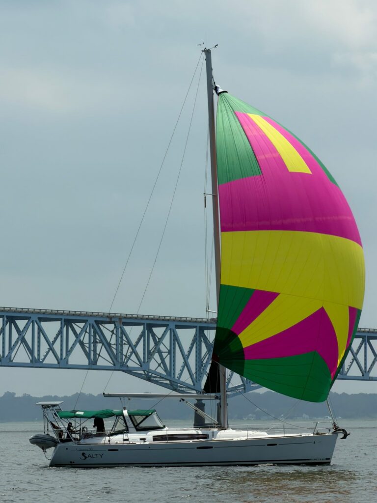 SV Salty sails under a big pink, green and yellow spinnaker on gray water, with a gray inflatable dinghy on the stern davits and a long steel truss bridge in the background under an overcast sky.