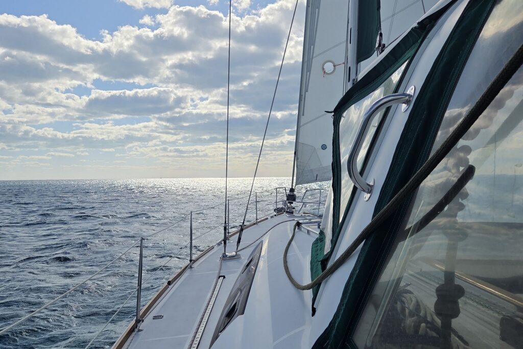 View forward along SV Salty's side deck under sail, with the white mainsail, green-trimmed dodger windows and lifelines beside open, sunlit ocean under scattered clouds.
