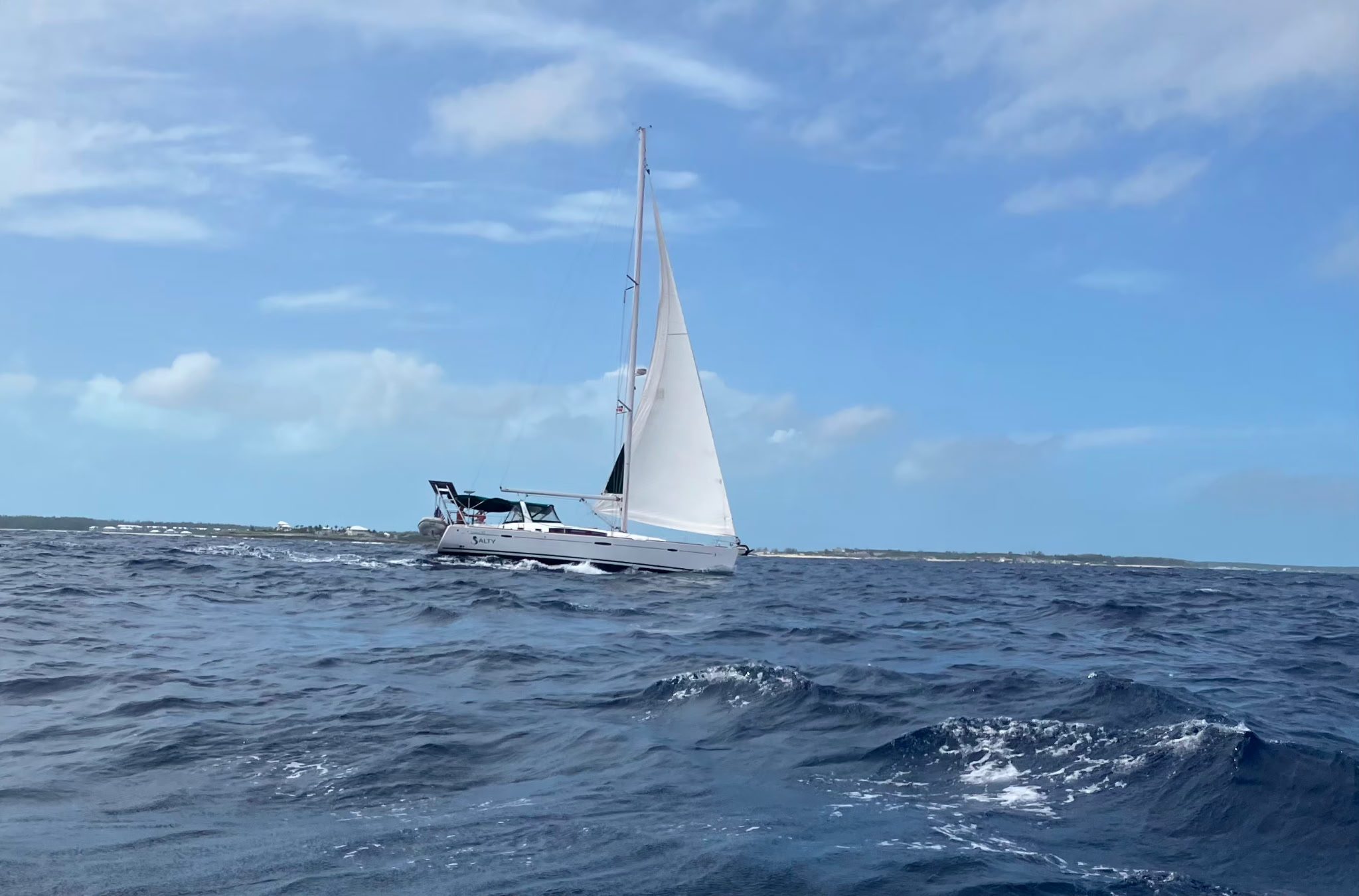 SV Salty heels slightly under a full white sail on choppy deep-blue water, seen from another boat, with a low island shoreline and a bright sky of scattered clouds behind her.
